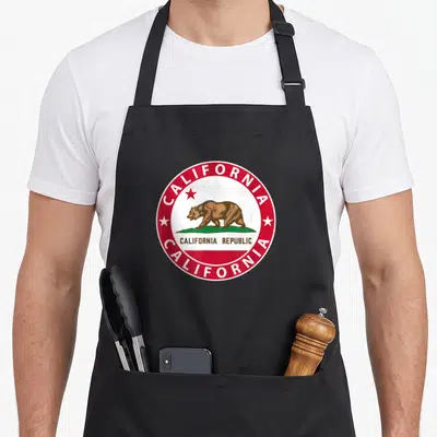 Tablier de cuisine noir avec le logo circulaire rouge et blanc du drapeau California représentant un ours brun sur une touffe d'herbe verte.