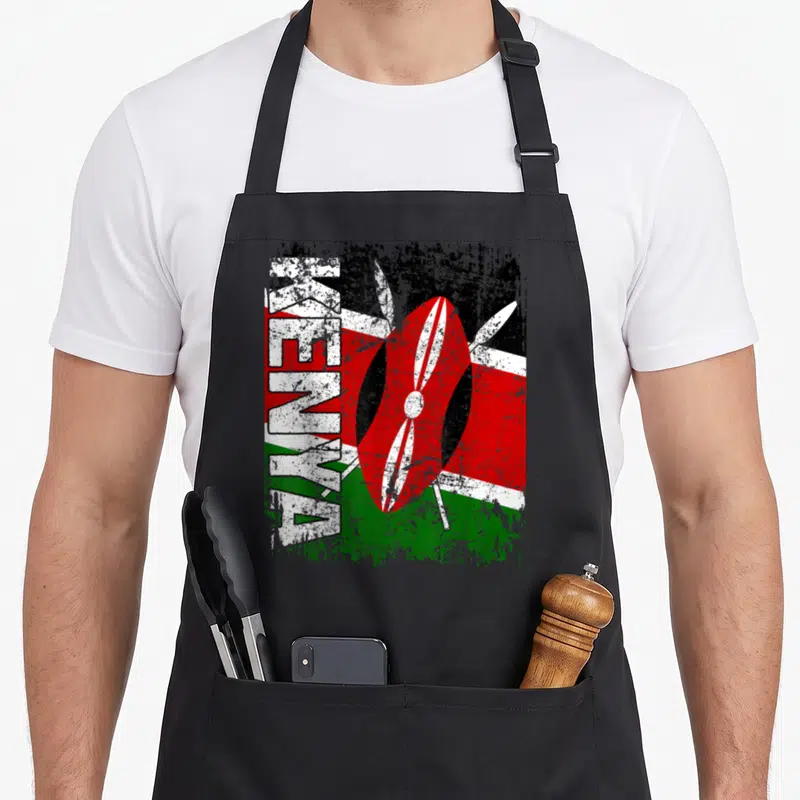 Tablier de cuisine noir avec inscription KENYA verticale, bouclier traditionnel rouge et lances blanches croisées sur fond aux couleurs du drapeau.