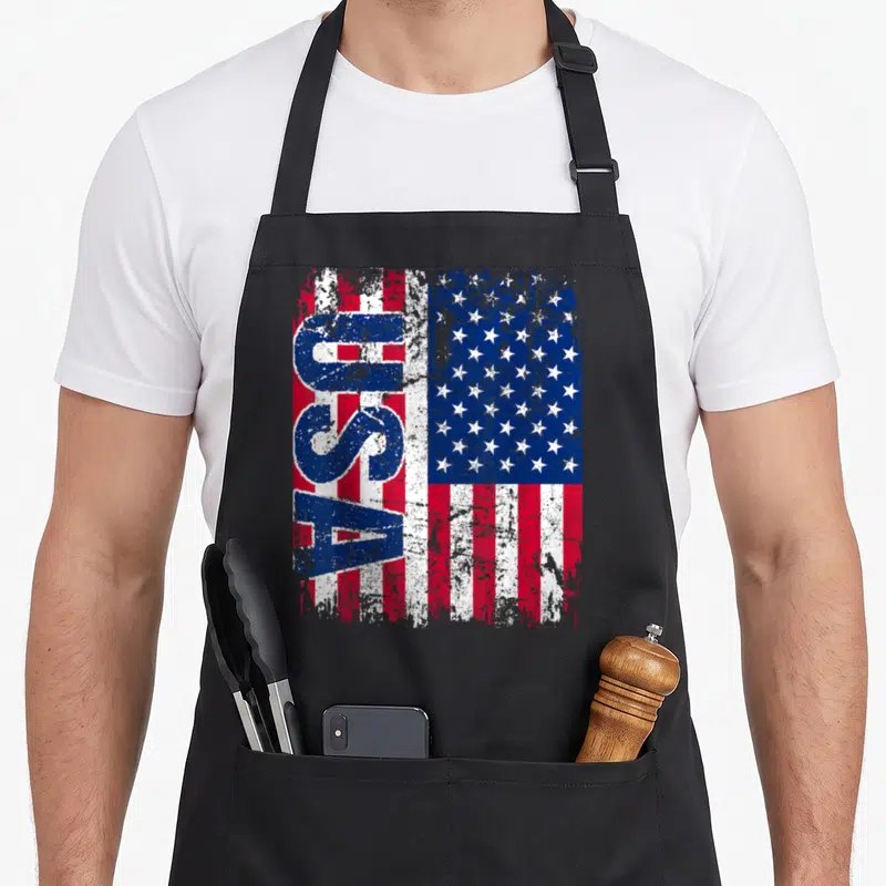 Tablier de cuisine noir avec le drapeau américain et l'inscription USA en lettres bleues verticales sur fond de bandes rouges et blanches.