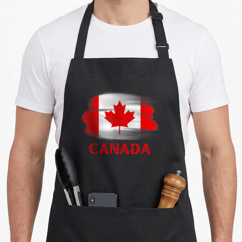 Tablier de cuisine noir avec un motif représentant le drapeau du Canada stylisé et l'inscription CANADA en rouge.