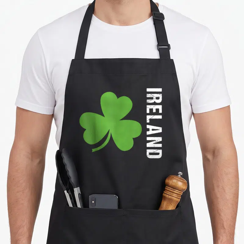 Tablier de cuisine noir avec grand trèfle vert et texte Ireland vertical blanc texturé sur le côté droit.