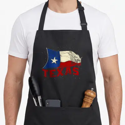 Tablier de cuisine noir avec un drapeau texan ondulé et le texte TEXAS en lettres rouges à effet pointilliste.