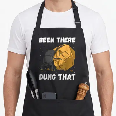 Tablier de cuisine noir avec illustration d'un bousier poussant une boule orange et le texte BEEN THERE DUNG THAT en blanc.