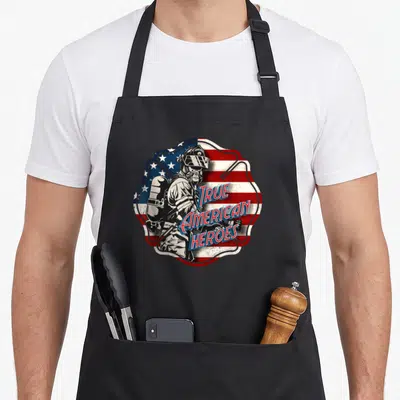 Tablier de cuisine noir avec illustration d'un pompier sur fond de drapeau américain et texte True American Heroes en lettres rouges et bleues.