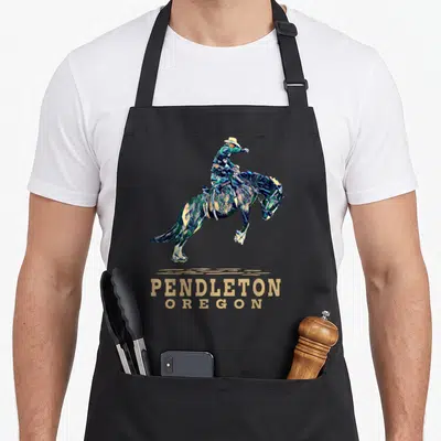 Tablier de cuisine noir avec illustration graphique d'un cowboy sur un cheval sauteur et le texte Pendleton Oregon en lettres dorées.