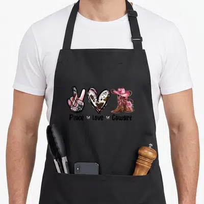 Tablier de cuisine noir avec main peace, cœur tacheté, bottes roses et chapeau country imprimés en couleur.