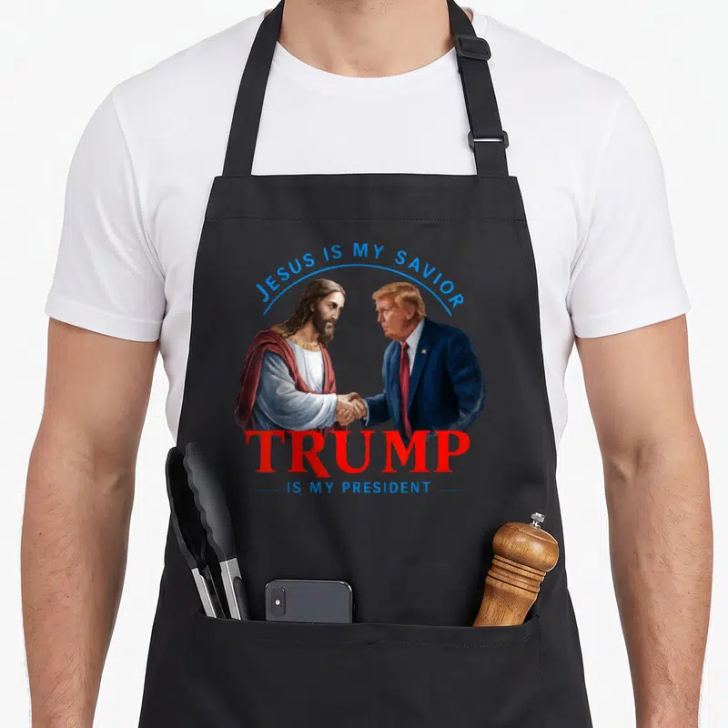 Tablier de cuisine noir avec illustration de Jésus et Donald Trump se serrant la main sous un drapeau américain stylisé.