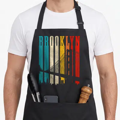 Tablier de cuisine noir avec le mot Brooklyn en lettres verticales multicolores stylisées en gratte-ciel et pont vintage.