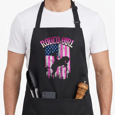 Tablier de cuisine noir avec silhouette cowgirl rodeo et texte Rodeo Girl sur drapeau américain dégradé rose et bleu.