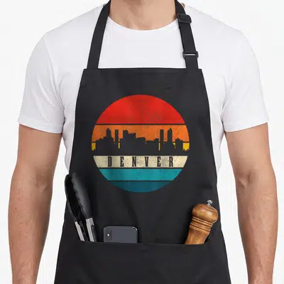 Tablier de cuisine noir avec un motif circulaire coloré représentant le skyline de Denver en style vintage.