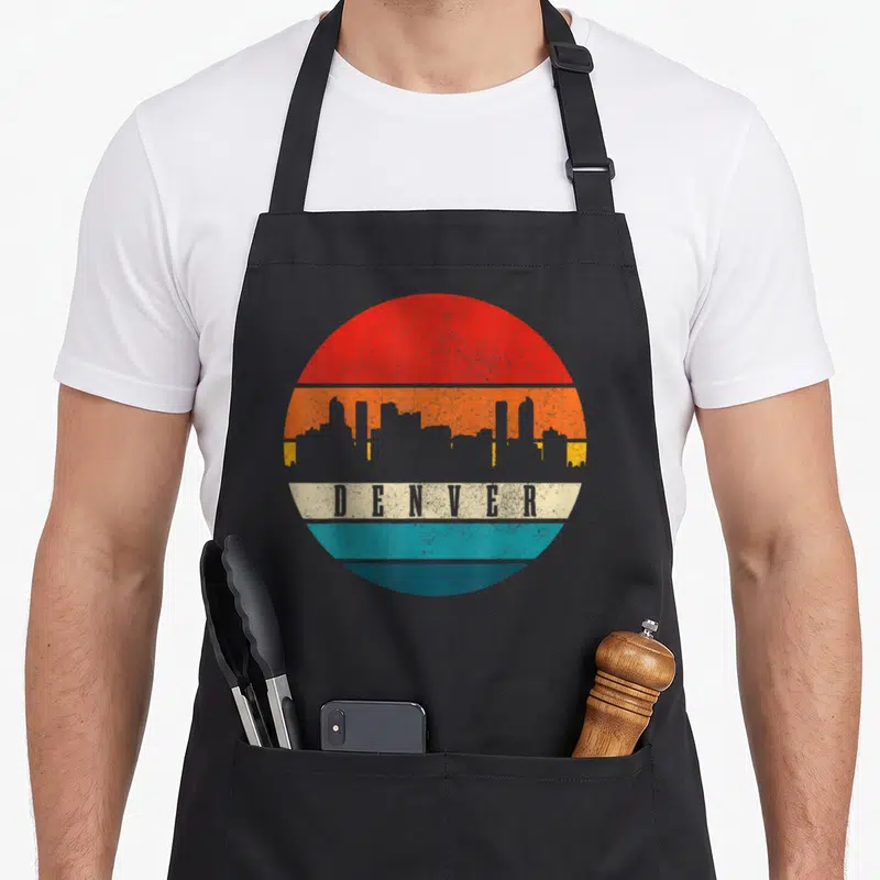 Tablier de cuisine noir avec un motif circulaire coloré représentant le skyline de Denver en style vintage.