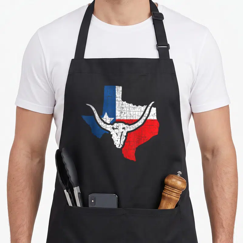 Tablier de cuisine noir avec une carte du Texas rouge blanche et bleue surmontée d'un crâne de taureau blanc.