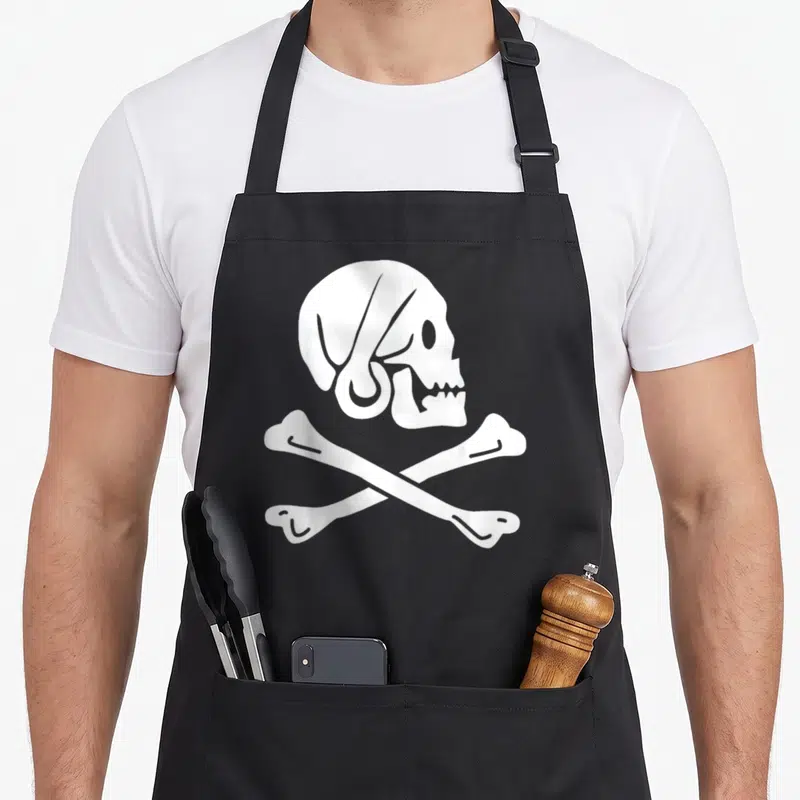 Tablier de cuisine noir avec un crâne blanc et deux os croisés en forme de Jolly Roger pirate.