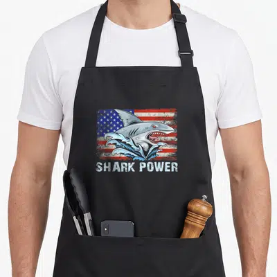 Tablier de cuisine noir avec un requin sortant de l'eau devant le drapeau américain et le texte Shark Power en blanc.