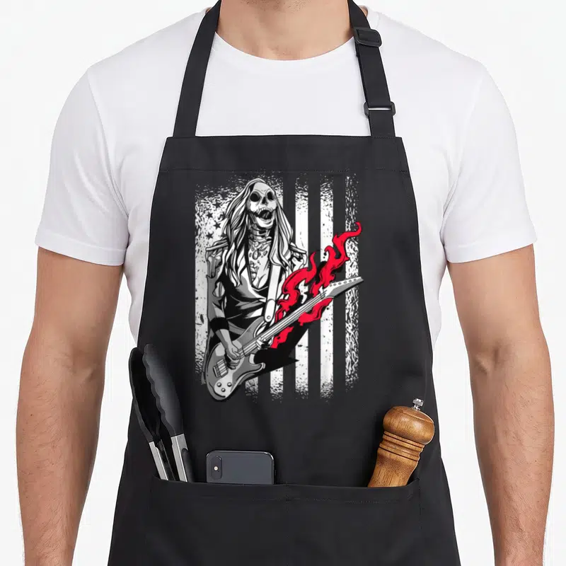 Tablier de cuisine noir avec une zombie guitariste au crâne visible et une guitare enflammée sur fond de drapeau USA.