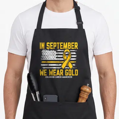 Tablier de cuisine noir avec un drapeau américain stylisé en blanc et or, surmonté d'un ruban jaune et du texte In September Nous Portons Or Childhood Cancer Awareness.