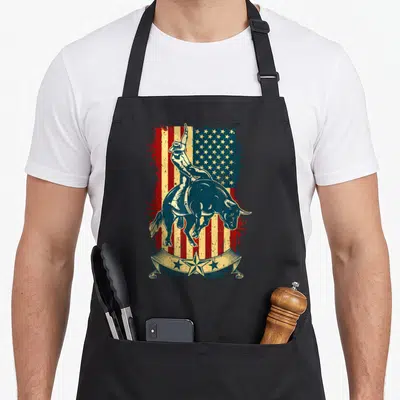 Tablier de cuisine noir avec illustration d'un cavalier de rodeo sur un taureau devant un drapeau américain vintage.