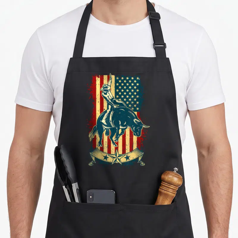 Tablier de cuisine noir avec illustration d'un cavalier de rodeo sur un taureau devant un drapeau américain vintage.