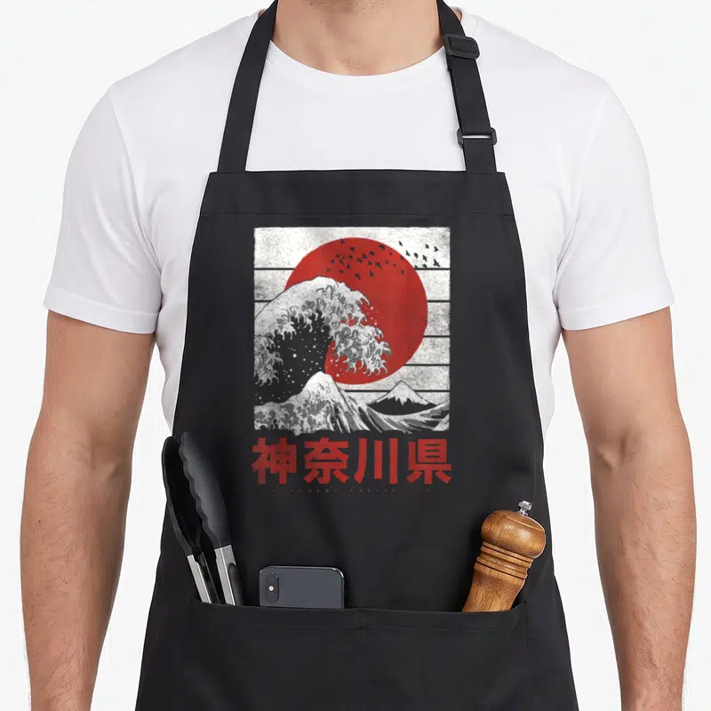 Tablier de cuisine noir avec une illustration stylisée d'une grande vague blanche, d'un soleil rouge et de caractères japonais rouges en bas.