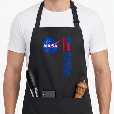 Tablier de cuisine noir avec le logo circulaire NASA, un drapeau américain et une inscription verticale bleue.
