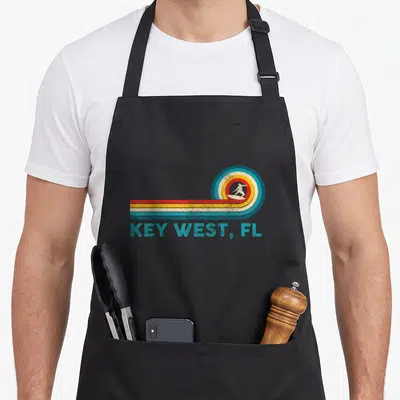 Tablier de cuisine noir avec illustration vintage d'un surfeur dans une vague multicolore et le texte KEY WEST, FL en bas.
