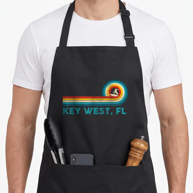 Tablier de cuisine noir avec illustration vintage d'un surfeur dans une vague multicolore et le texte KEY WEST, FL en bas.