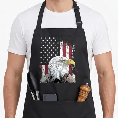 Tablier de cuisine noir avec tête d'aigle blanc et drapeau américain délavé en arrière-plan.