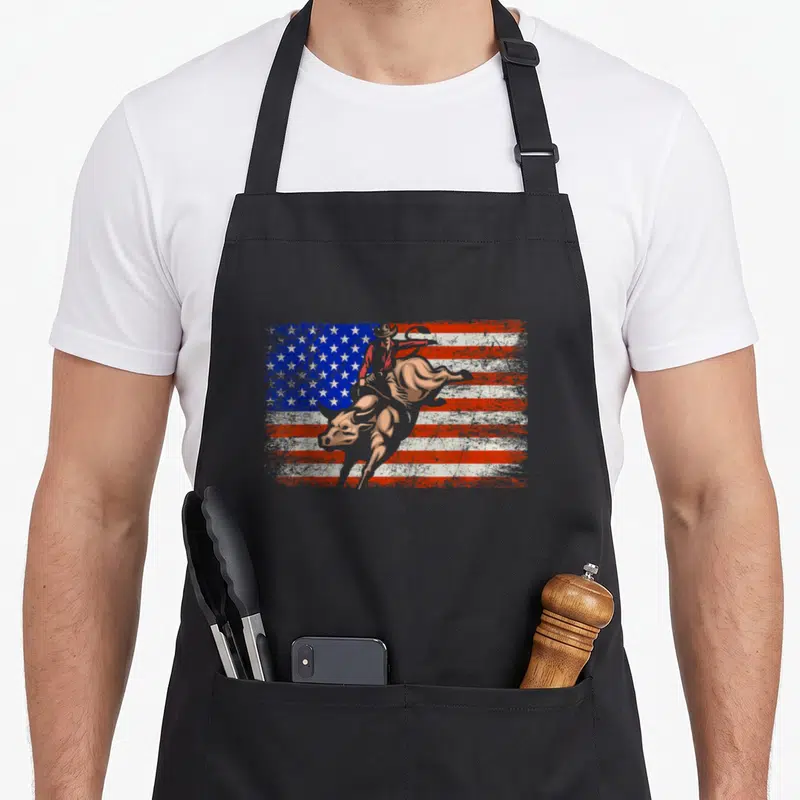 Tablier de cuisine noir avec illustration d'un cowboy sur un taureau devant un drapeau américain texturé.