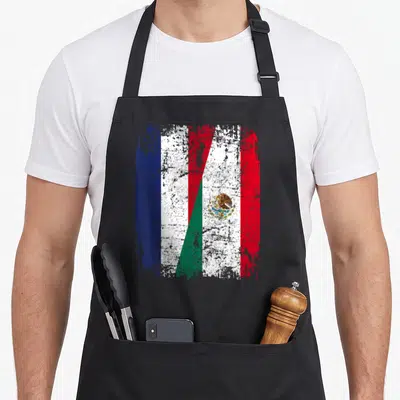 Tablier de cuisine noir avec un drapeau stylisé fusionnant les couleurs françaises et mexicaines sur fond texturé.