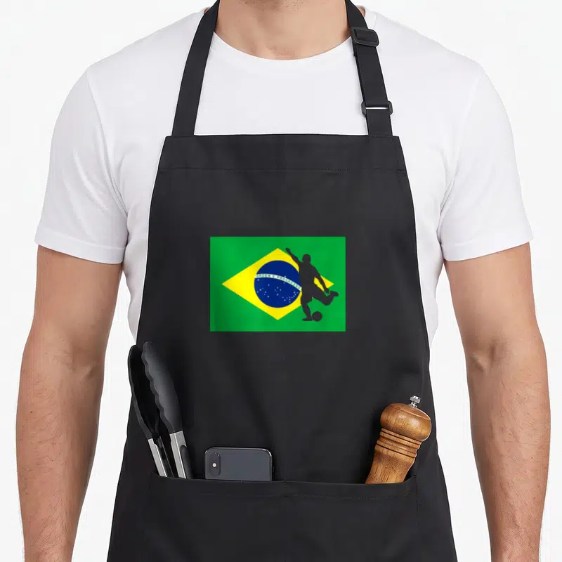 Tablier de cuisine noir avec drapeau Brésil Brasil et silhouette grise d'un joueur de football en action sur fond vert et jaune.