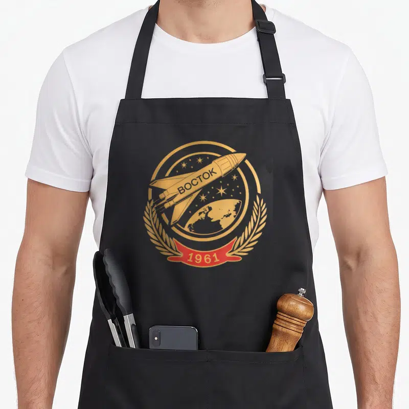 Tablier de cuisine noir avec logo doré Vostok 1961 et fusée spatiale soviétique.