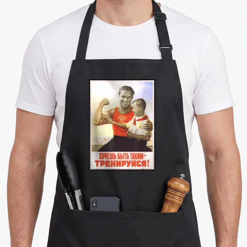 Tablier de cuisine noir avec une affiche soviétique montrant un homme musclé tenant un enfant et du texte en russe rouge en bas.