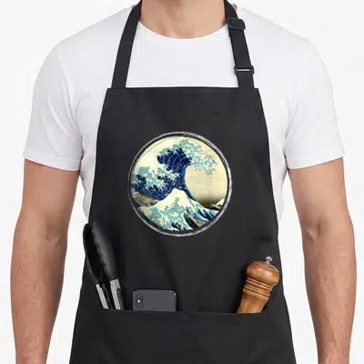 Tablier de cuisine noir avec une grande vague stylisée et des bateaux imprimés en cercle au centre.