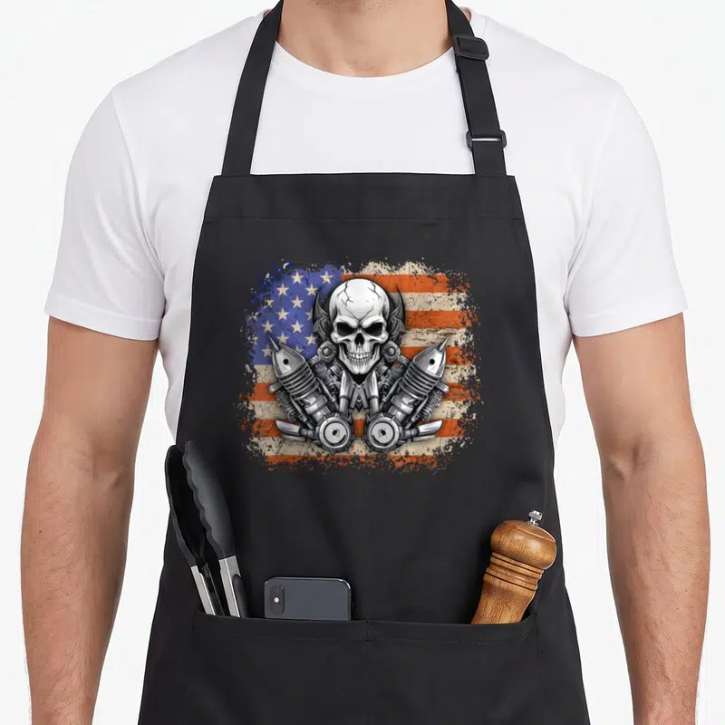 Tablier de cuisine noir avec un crâne de motard sur drapeau américain et pièces de moteur métalliques.