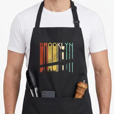 Tablier de cuisine noir avec inscription BROOKLYN colorée et silhouette du pont de Brooklyn intégrée au design graphique.