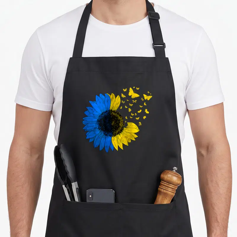 Tablier de cuisine noir avec un grand tournesol bleu et jaune dont les fleurs s'envolent sous forme de papillons dorés.
