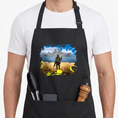 Tablier de cuisine noir avec illustration d'un soldat face à un navire de guerre sous des couleurs bleue et jaune.