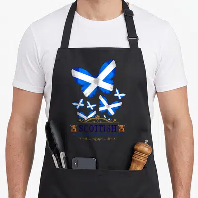 Tablier de cuisine noir avec des papillons au drapeau écossais, le mot Scottish et deux têtes de bœuf highland sur les côtés.