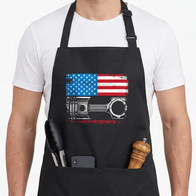 Tablier de cuisine noir avec drapeau américain vintage et illustration de piston mécanique en blanc.