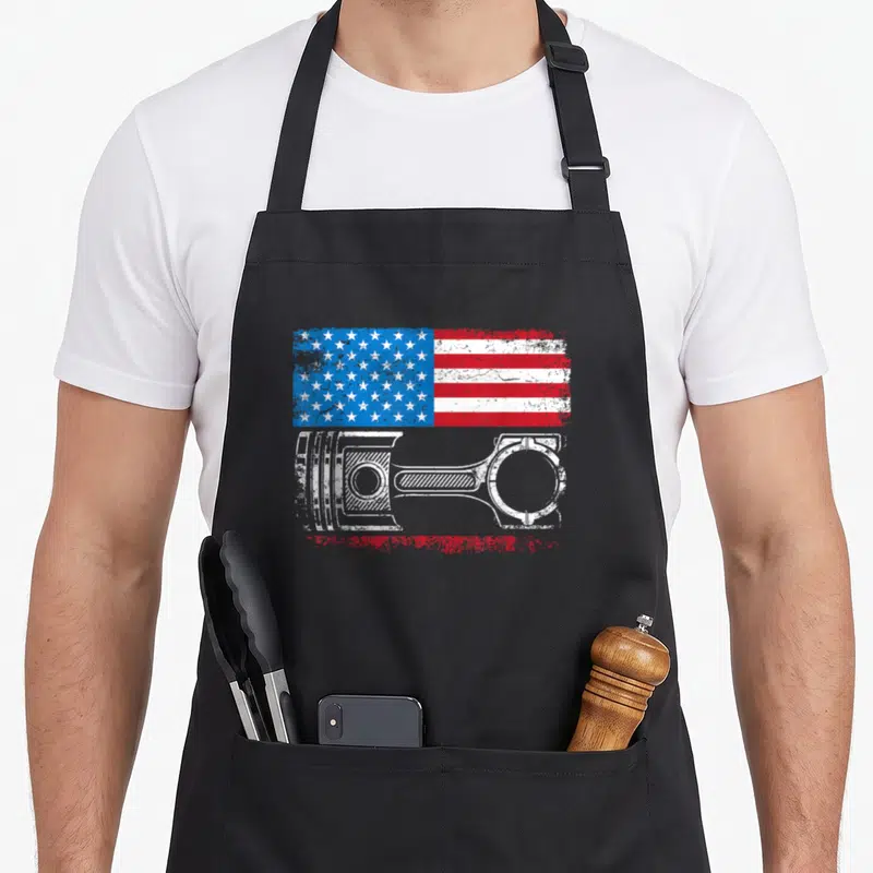 Tablier de cuisine noir avec drapeau américain vintage et illustration de piston mécanique en blanc.