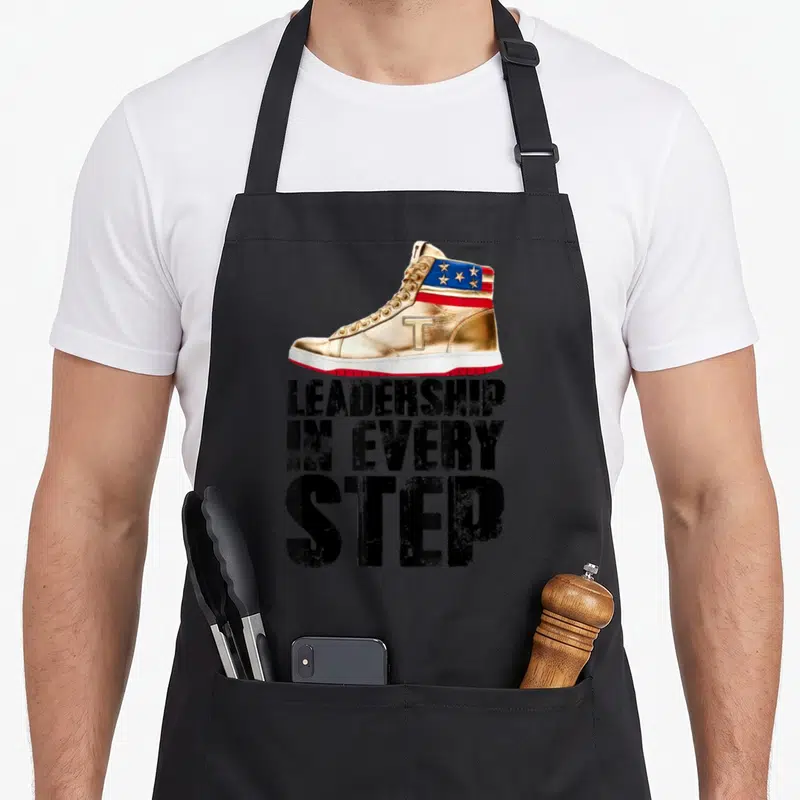 Tablier de cuisine noir avec une illustration de sneaker dorée et le texte LEADERSHIP IN EVERY STEP en lettres noires capitales.