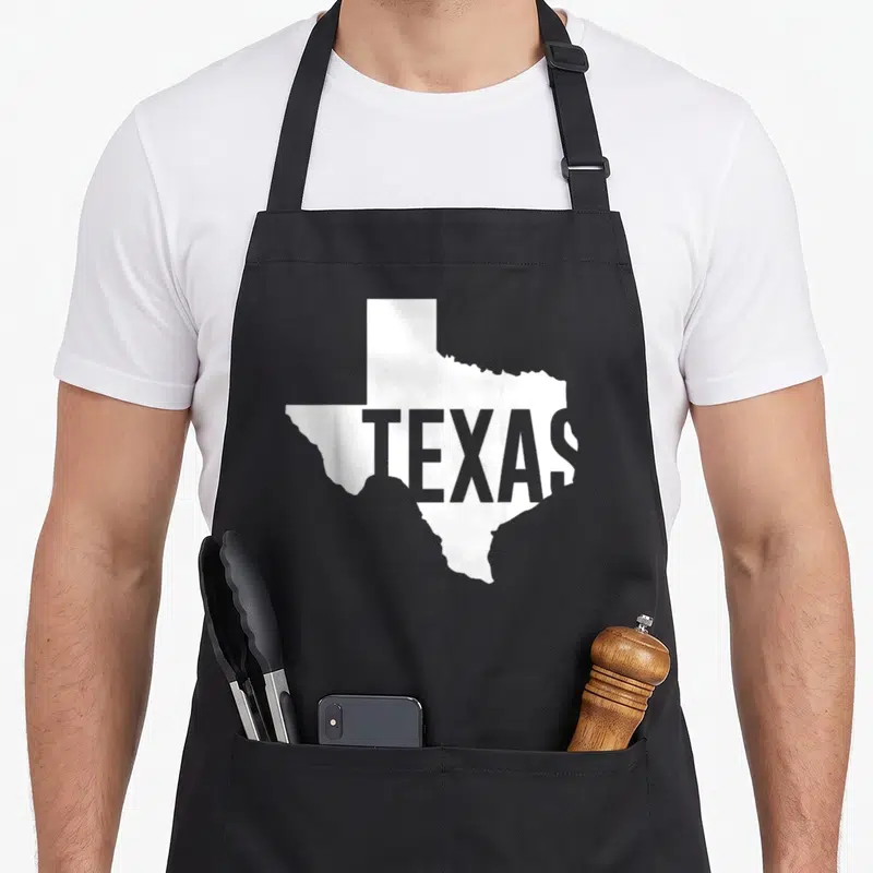 Tablier de cuisine noir avec silhouette blanche du Texas et le mot TEXAS en gris foncé au centre du motif.