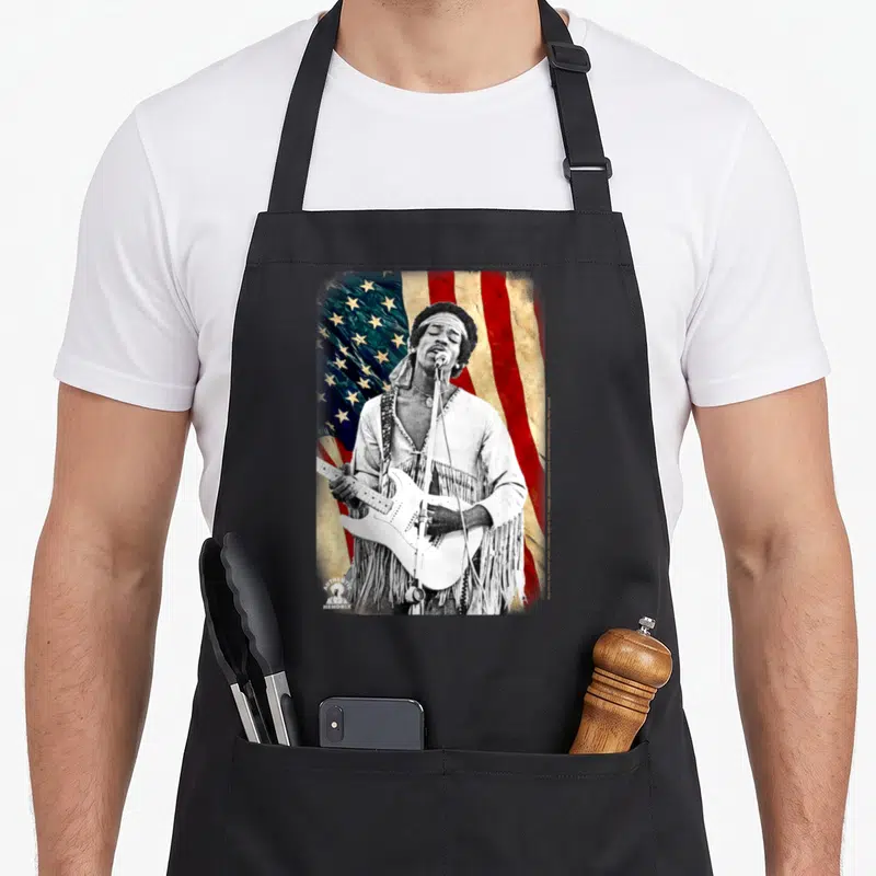 Tablier de cuisine noir avec une illustration vintage de Jimi Hendrix jouant devant une American Flag et le logo Hendrix en bas à gauche.