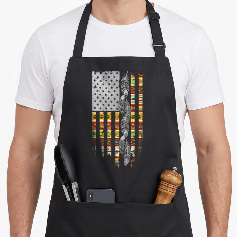 Tablier de cuisine noir avec un drapeau amérindien stylisé, des plumes grises et des franges aux motifs tribaux multicolores.