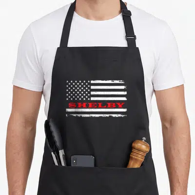 Tablier de cuisine noir avec un drapeau américain stylisé et le mot Shelby en lettres rouges au centre.