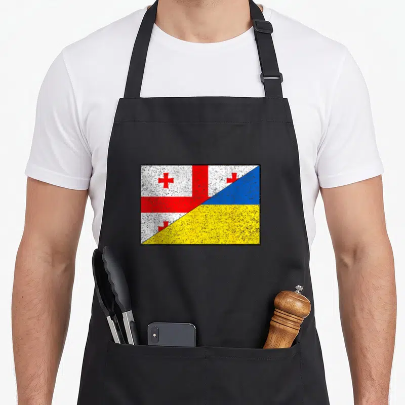 Tablier de cuisine noir avec un drapeau divisé en deux montrant la Géorgie et l'Ukraine sur fond texturé.