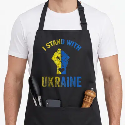 Tablier de cuisine noir avec un poing aux couleurs du drapeau ukrainien et un texte de soutien imprimé en jaune et bleu.