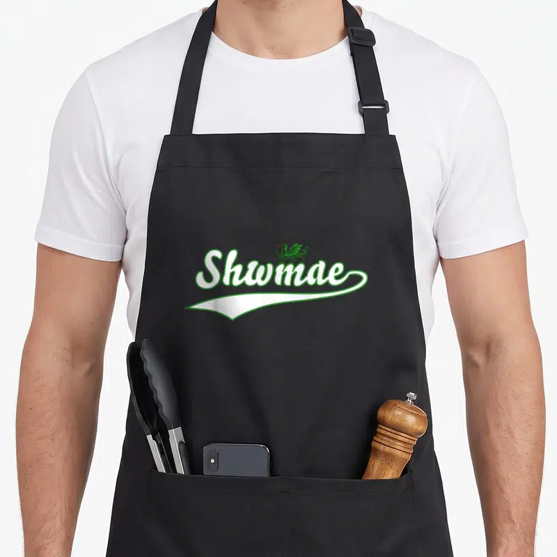 Tablier de cuisine noir avec l'inscription Shwmae et un dragon vert posé sur la lettre e.