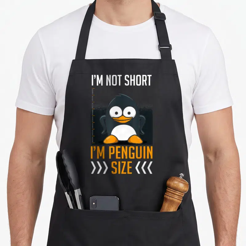 Tablier de cuisine noir avec un pingouin souriant, une règle et le texte I'm Not Short I'm Penguin Size imprimé en blanc et orange.