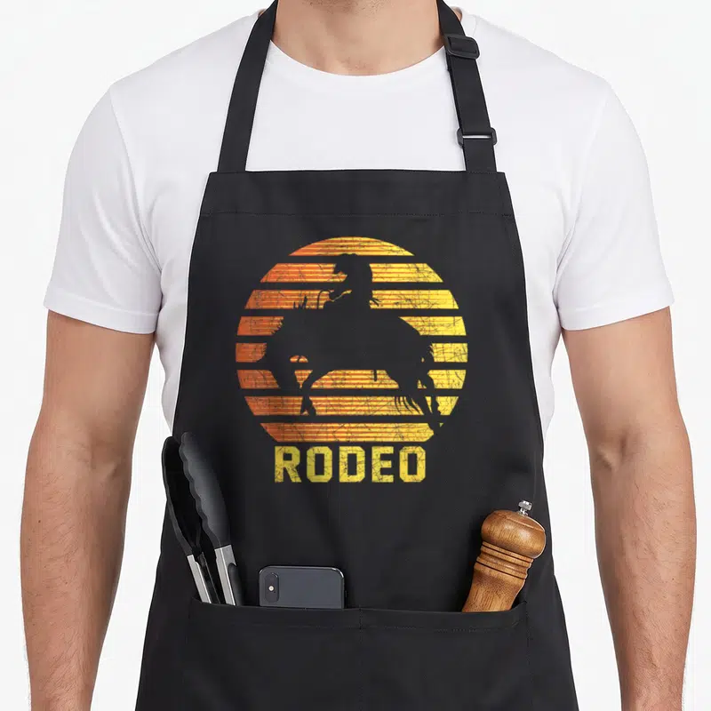 Tablier de cuisine noir avec silhouette de cheval de rodeo et texte RODEO sur fond de soleil rayé orange et jaune.
